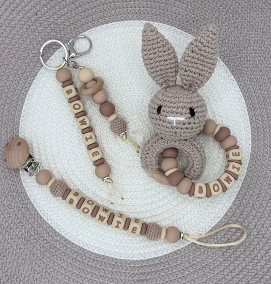 A baby accessory set featuring a beige crochet bunny teething ring with a wooden bead ring, a matching wooden letter clip, and a wooden bead clip, all arranged on a white circular mat inside a large taupe woven hat.